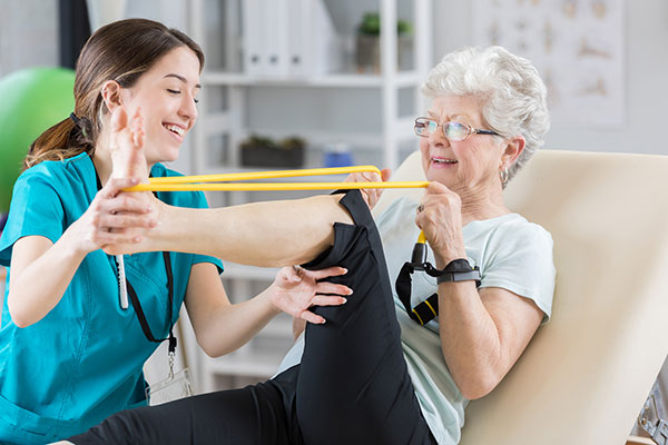 Confident Caucasian female physical therapist helps senior Caucasian patient use resistance band.